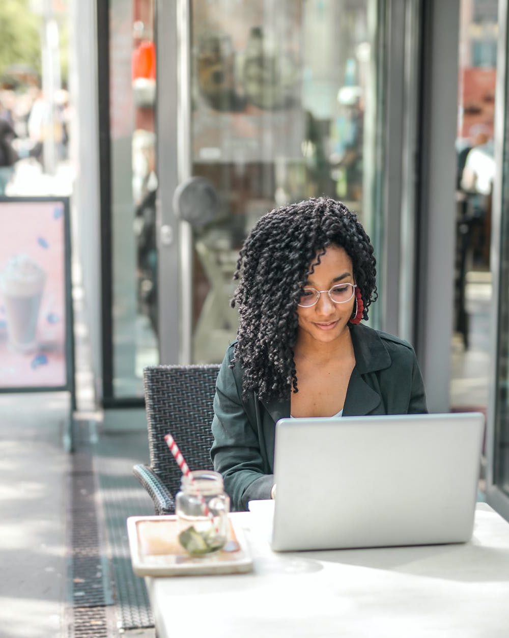 ethnic young woman using laptop while having tasty beverage in modern street cafe